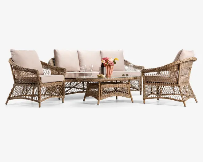 Sættet har et flettet rattan-design i en naturlig brun farve og består af en trepersoners sofa, to lænestole og et rektangulært sofabord med glasplade. Siddepladserne har tykke, lyse beige hynder på både sæde og ryg for ekstra komfort. Armlæn og baser har et åbent gittermønster, som giver et let og elegant udtryk. Sofabordet er pyntet med en vase med blomster, en flaske, glas og en lille tallerken, hvilket understreger dets anvendelse til servering.