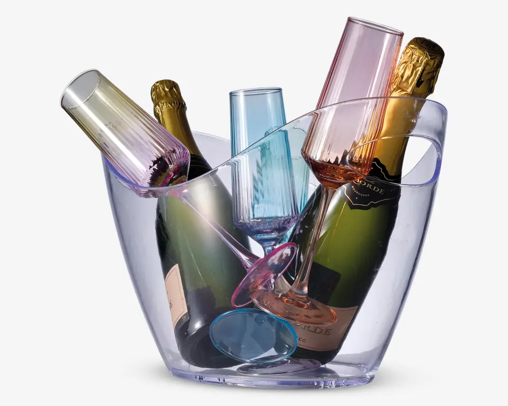 En gennemsigtig, oval isspand indeholder to flasker mousserende vin og fire farvede champagneglas. Glassene er lavet af tonet glas i pastelfarverne pink, blå, grøn og lilla og er placeret i forskellige vinkler mellem flaskerne. Spanden har et glat, moderne design med let udadbuede sider.