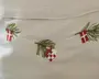 Billedet viser et nærbillede af broderi på et lyst stof. Motivet forestiller en snor med små, detaljerede ophæng, herunder grønne grangrene, røde og hvide dannebrogsflag samt et rødt og hvidt flettet julehjerte. Broderiet er udført med fine sting og giver et festligt og traditionelt skandinavisk juleudtryk.