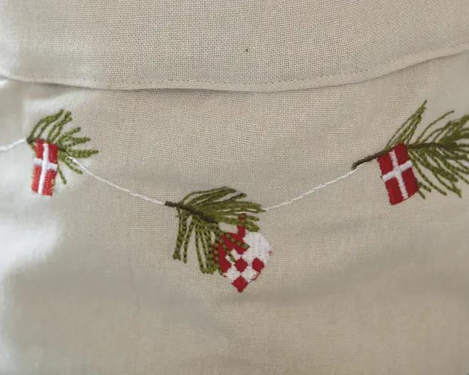 Billedet viser et nærbillede af broderi på et lyst stof. Motivet forestiller en snor med små, detaljerede ophæng, herunder grønne grangrene, røde og hvide dannebrogsflag samt et rødt og hvidt flettet julehjerte. Broderiet er udført med fine sting og giver et festligt og traditionelt skandinavisk juleudtryk.
