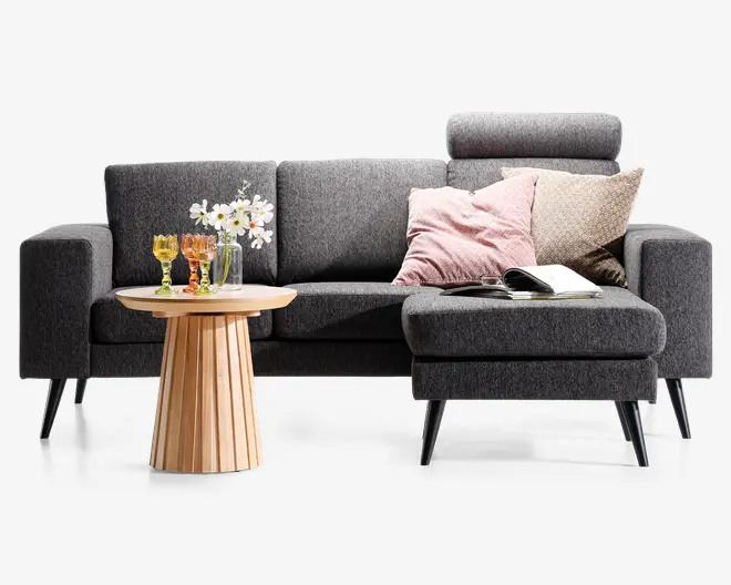 Moderne L-formet sofa betrukket med mørkegråt stof og slanke, skrå sorte ben. Sofaen har en chaiselong til højre, et nakkestøtte på et af sæderne og er pyntet med to pyntepuder – én lyserød og én beige med mønster. En åben bog ligger på chaiselongen. Foran sofaen står et rundt sidebord i træ med rillet fod, hvorpå der står en vase med hvide blomster og tre farvede glas.