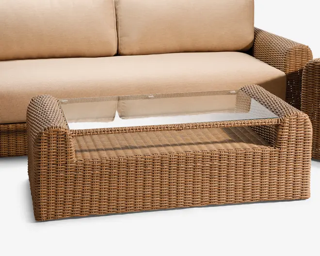 Rektangulært sofabord med en ramme i flettet rattan-look i lysebrun farve. Bordet har en indsat, klar glasplade, som understøttes af rammens flet. Designet har afrundede hjørner og en lavere hylde under glaspladen til ekstra opbevaring eller udstilling.