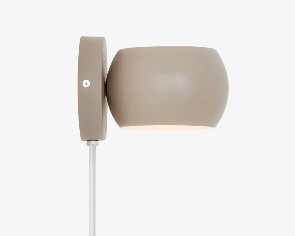 En moderne vægmonteret lampe med en glat, mat taupe-farvet overflade. Lampen har en ovalt formet skærm, der leder lyset nedad, samt en rektangulær vægplade med en synlig skrue til montering. En hvid ledning går ud fra bunden af vægpladen. Designet er minimalistisk og moderne, velegnet til forskellige indretninger.