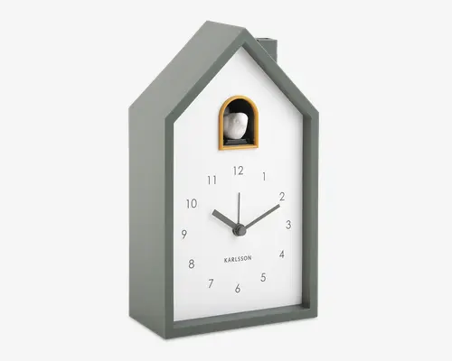 Moderne ur med husformet ramme i mat grå, hvid urskive med sorte arabertal og minimalistiske sorte visere. Øverst er der et lille buet vindue med gul kant, hvor en dekorativ fuglefigur ses, hvilket minder om et traditionelt kukur. Mærket 'Karlsson' står trykt over klokken 6.