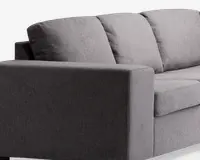 Nærbillede af en moderne sofa med minimalistisk design og rene linjer. Betrækket er lavet af et tekstureret, mellemgråt stof. Armlænet er bredt og rektangulært, og ryghynderne er bløde og let skrånende for komfort. Sædehynderne er tykke og faste, hvilket giver god støtte.