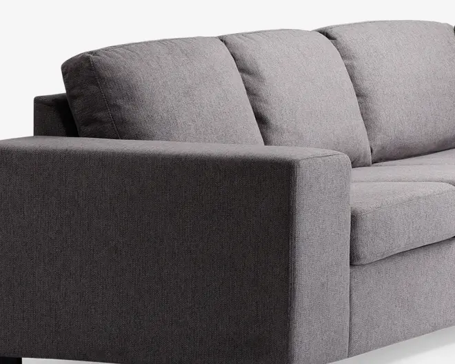 Nærbillede af en moderne sofa med minimalistisk design og rene linjer. Betrækket er lavet af et tekstureret, mellemgråt stof. Armlænet er bredt og rektangulært, og ryghynderne er bløde og let skrånende for komfort. Sædehynderne er tykke og faste, hvilket giver god støtte.