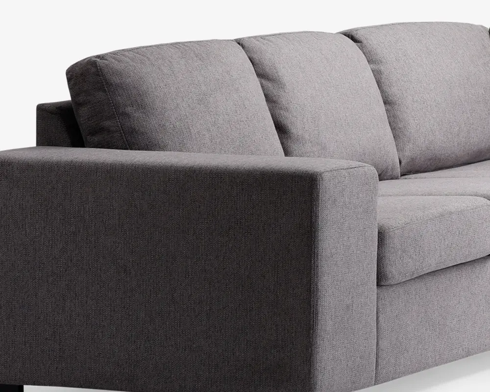 Nærbillede af en moderne sofa med minimalistisk design og rene linjer. Betrækket er lavet af et tekstureret, mellemgråt stof. Armlænet er bredt og rektangulært, og ryghynderne er bløde og let skrånende for komfort. Sædehynderne er tykke og faste, hvilket giver god støtte.