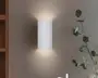 En moderne vægmonteret lampe med en lodret rillet, cylindrisk hvid skærm. Lyset udsendes blødt og diffust både opad og nedad, hvilket skaber en stemningsfuld effekt på væggen. Lampen har et minimalistisk design, der passer til moderne indretning.
