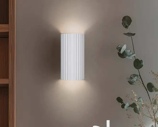 En moderne vægmonteret lampe med en lodret rillet, cylindrisk hvid skærm. Lyset udsendes blødt og diffust både opad og nedad, hvilket skaber en stemningsfuld effekt på væggen. Lampen har et minimalistisk design, der passer til moderne indretning.
