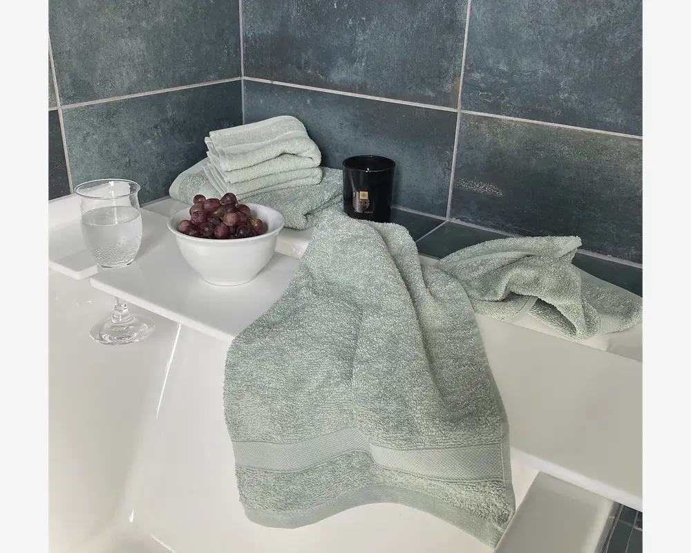 Bløde, lysegrønne håndklæder med en diskret kantdetalje, præsenteret i et badeværelsesmiljø. Håndklæderne ser tykke og absorberende ud med en blid tekstur, der er velegnet til sensitiv hud. De vises i forskellige størrelser, pænt foldet og lagt, hvilket fremhæver deres kvalitet og fyldighed.