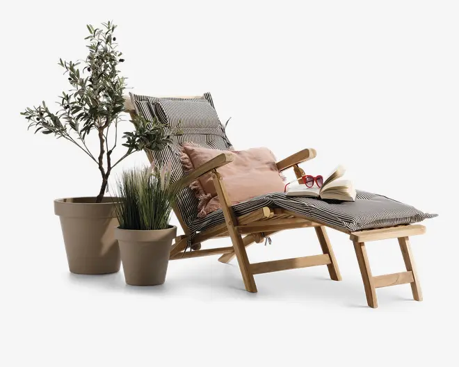 En liggestol i træ med tilhørende fodskammel, begge med sort-hvidstribede hynder. Stolen har en blød, beige pyntepude, et par røde læsebriller og en åben bog placeret på fodskamlen. Ved siden af stolen står to potteplanter: én høj med slanke blade og én lavere med græslignende blade, hvilket giver et grønt og hyggeligt udtryk.
