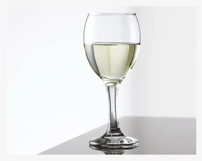 Et klart glas med stilk og rund skål, delvist fyldt med en lysegul væske. Glasset har en glat, let udadbuet kant og en solid fod, velegnet til servering af hvidvin eller lignende drikke. Overfladen er reflekterende, og glasset fremstår rent og gennemsigtigt.