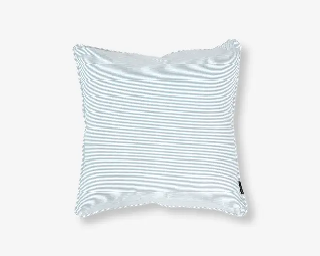 Firkantet pude med et diskret, fint lyseblåt og hvidt nålestribet mønster. Stoffet ser blødt og glat ud, og puden har et let overfyldt, fyldigt udseende. Der er et lille sort stofmærke på den ene kant, sandsynligvis med mærkets navn. Kanterne er pænt afsluttede med en let piping-detalje.