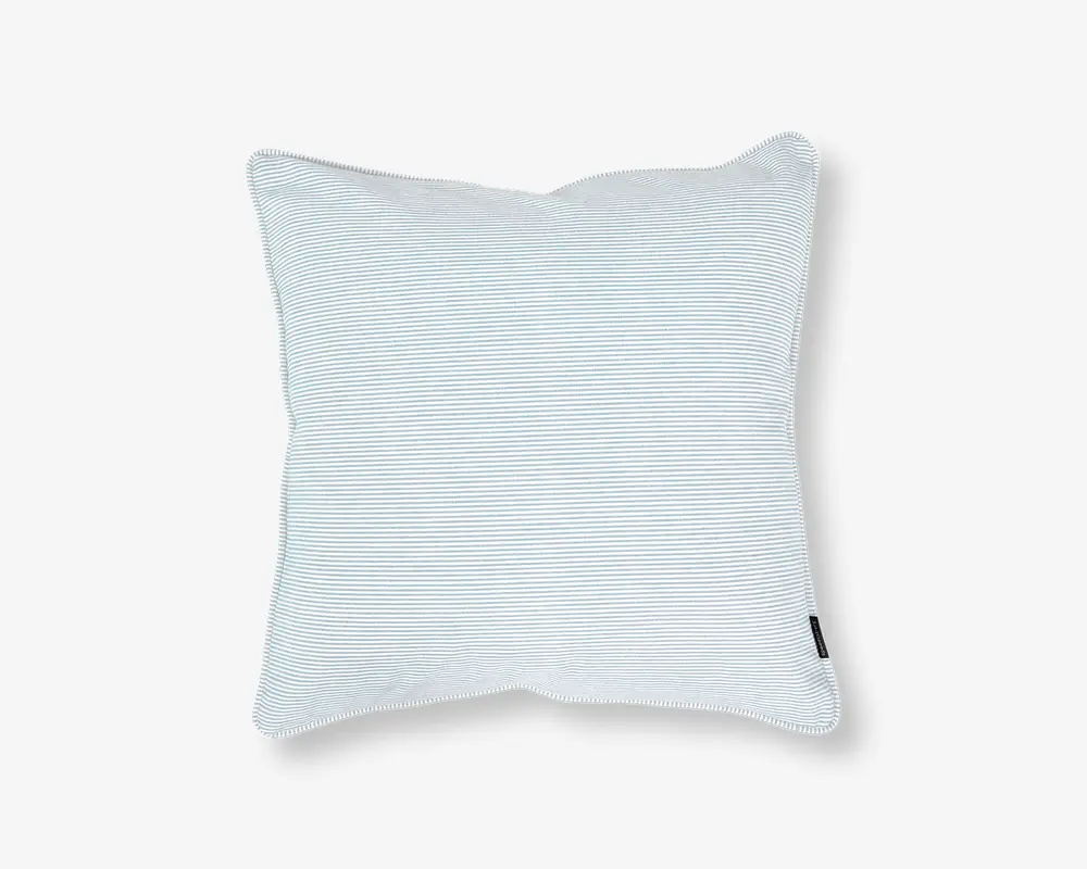 Firkantet pude med et diskret, fint lyseblåt og hvidt nålestribet mønster. Stoffet ser blødt og glat ud, og puden har et let overfyldt, fyldigt udseende. Der er et lille sort stofmærke på den ene kant, sandsynligvis med mærkets navn. Kanterne er pænt afsluttede med en let piping-detalje.