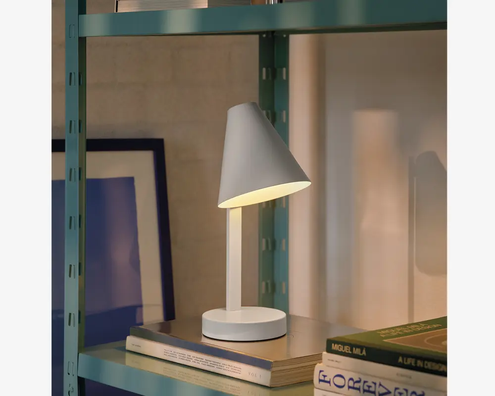 En moderne bordlampe med mat hvid finish, der har en konisk skærm vinklet nedad for fokuseret belysning. Lampen har en slank, lige stang og en rund, stabil fod. Den er kompakt i størrelsen, velegnet til placering på hylder eller skriveborde, og giver et blødt, varmt lys, der er ideelt til læsning eller stemningsbelysning.
