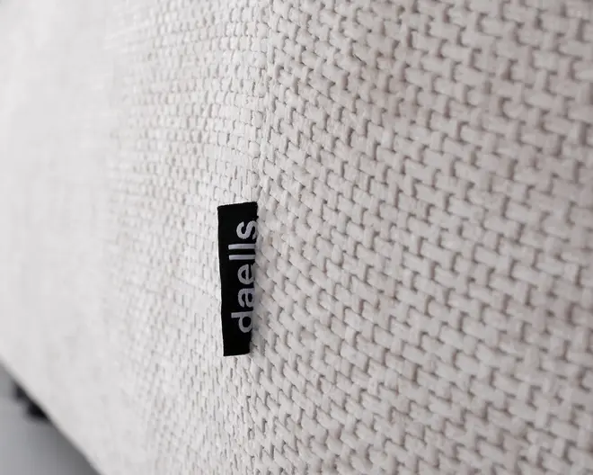 Nærbillede af et lyst beige, vævet stof med tydelig tekstur. Et lille sort stofmærke med mærket 'daells' i hvid tekst er syet ind i sømmen. Vævemønstret er tæt og ensartet, hvilket giver overfladen et slidstærkt og let tekstureret udseende.