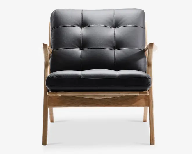 Stol med sort, polstret pude med synlige syninger og knapper, der giver et blødt og komfortabelt sæde og ryglæn. Stellet er lavet af lyst træ med en naturlig finish og har skrå ben og armlæn for et moderne, minimalistisk udtryk. Sæde- og ryghynderne er tykke og velpolstrede, hvilket giver god ergonomisk støtte.