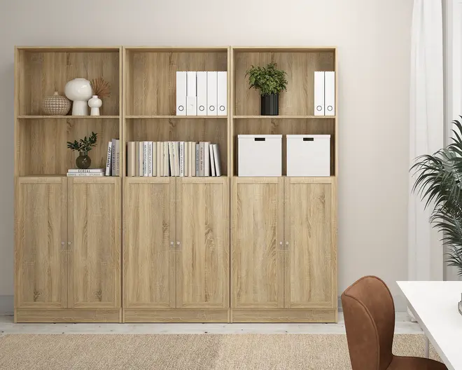 Tre høje reoler i lyst træ med naturlig træstruktur. Hver reol har to åbne hylder øverst og to lukkede skabslåger nederst. Hylderne er organiseret med hvide opbevaringskasser, hvide ringbind, bøger, dekorative genstande som en lampe, vaser og en lille potteplante. Skabslågerne har små runde greb. Designet er moderne og minimalistisk, velegnet til hjemmet eller kontoret.