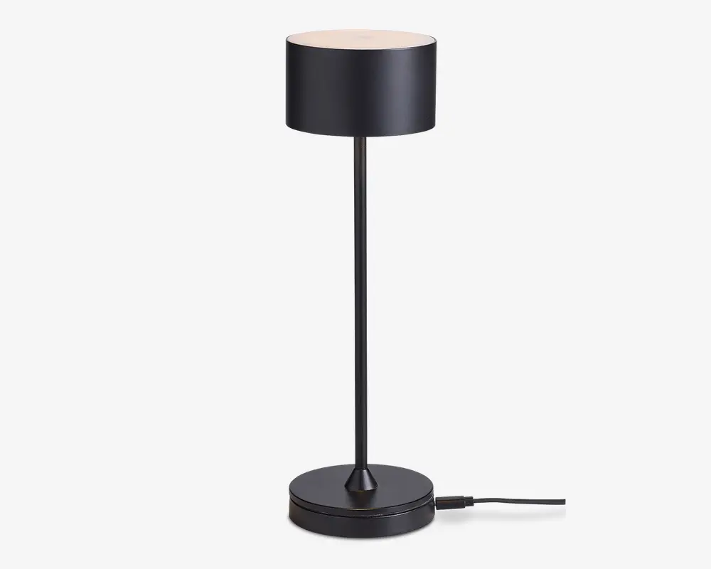En moderne bordlampe med en mat sort cylindrisk lampeskærm, slank lodret stang og rund fod. Lampen har et minimalistisk design med rene linjer og en glat overflade. Strømkablet går ud fra lampens fod, og skærmen spreder lyset jævnt fra toppen.