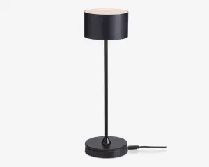 En moderne bordlampe med en mat sort cylindrisk lampeskærm, slank lodret stang og rund fod. Lampen har et minimalistisk design med rene linjer og en glat overflade. Strømkablet går ud fra lampens fod, og skærmen spreder lyset jævnt fra toppen.