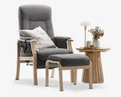 En moderne loungestol med tilhørende fodskammel, begge med lyse trærammer og polstret med blødt, mørkegråt stof. Stolen har høj ryg og polstrede armlæn for ekstra komfort. En stribet pude ligger på sædet. Ved siden af stolen står et rundt sidebord i træ med rillet fod, hvorpå der står en lille hvid lampe, en vase med tørrede blomster og et glas med snacks.