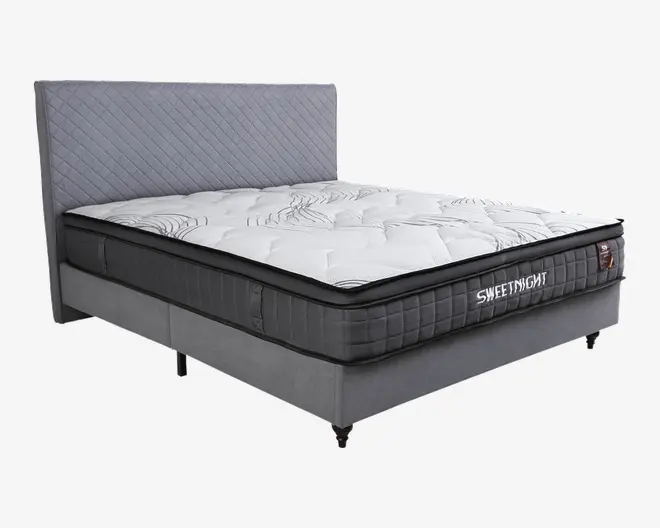 Moderne seng med høj, quiltet grå sengegavl og en tyk madras med hvid top og diskrete bølgemønstre. Madrassen har sort sidepanel med gitterstruktur og mærket 'SWEETNIGHT' broderet på siden. Sengebunden er polstret med gråt stof og står på metalben.