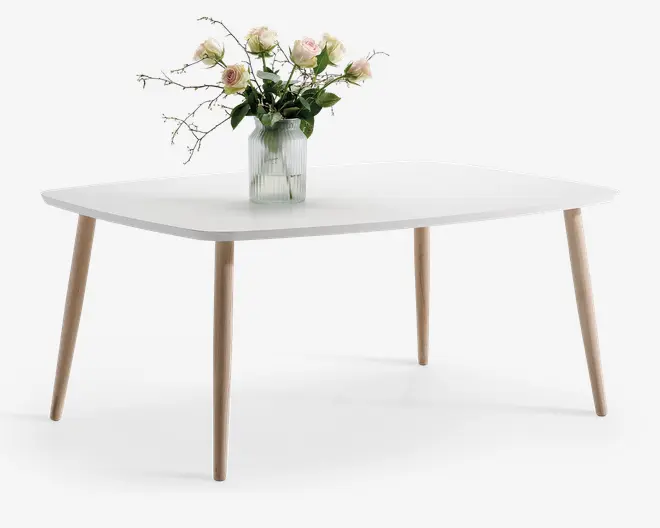 Rektangulært bord med en glat, mat hvid bordplade og fire skråtstillede ben i lyst træ. Bordet har et minimalistisk, moderne design. Midt på bordet står en rillet glasvase med en buket lyserøde og cremefarvede roser samt grønt løv.