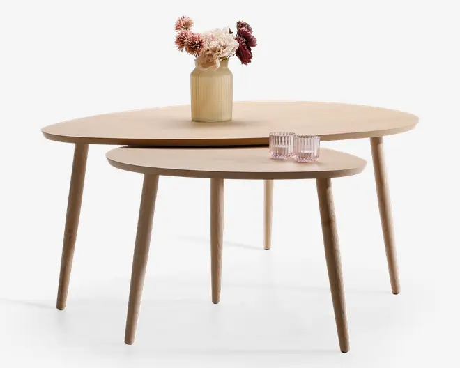 Sæt med to indskudsborde i lyst træ med runde bordplader. Bordene har slanke, koniske ben og et glat, minimalistisk design. Det større bord er pyntet med en rillet vase med kunstige blomster, mens det mindre bord har to lyserøde glas.