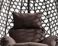 Nærbillede af en polstret siddeplads med en stor, blød rygpude og armlænspuder, alle i mørkebrunt stof. Sædet er placeret i en ramme med flettet, geometrisk mønster lavet af syntetisk rattan, hvilket giver et moderne og luftigt udtryk. Pudernes fyldighed og blødhed indikerer god komfort til afslapning.