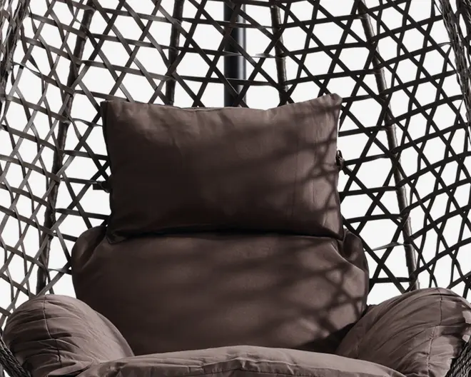 Nærbillede af en polstret siddeplads med en stor, blød rygpude og armlænspuder, alle i mørkebrunt stof. Sædet er placeret i en ramme med flettet, geometrisk mønster lavet af syntetisk rattan, hvilket giver et moderne og luftigt udtryk. Pudernes fyldighed og blødhed indikerer god komfort til afslapning.