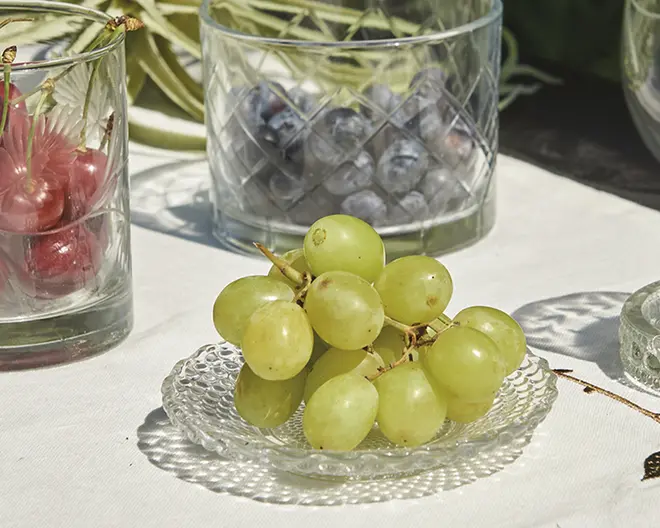 En lille, klar glasasiet rummer en klase friske grønne druer. Druerne er fyldige og blanke med et naturligt, let gennemsigtigt udseende. I baggrunden står to glas med slibemønster, det ene indeholder røde kirsebær og det andet blåbær. Scenen er sat på en hvid dug, hvilket giver et rent og elegant udtryk.