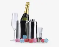 Billedet viser et sæt barudstyr, herunder en champagnekøler i rustfrit stål med en flaske champagne, en matchende cocktailshaker med låg, et klart champagneglas, et klart tumblerglas og flere genanvendelige isterninger i pink, blå og røde farver. Produkterne er arrangeret sammen og fremhæver det blanke metal og de farverige isterninger.