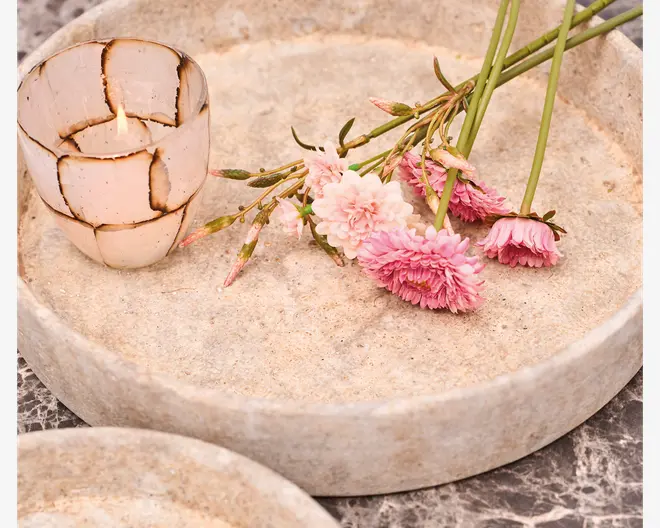 Rund, lav bakke lavet af lyse sten med en glat, mat overflade. Bakken har en forhøjet kant og viser en lille, dekorativ lysestage med et krakeleret, mosaiklignende mønster i hvide og brune nuancer. Der ligger også flere lyserøde og hvide blomster med grønne stilke på bakken, hvilket giver et blødt, naturligt udtryk.