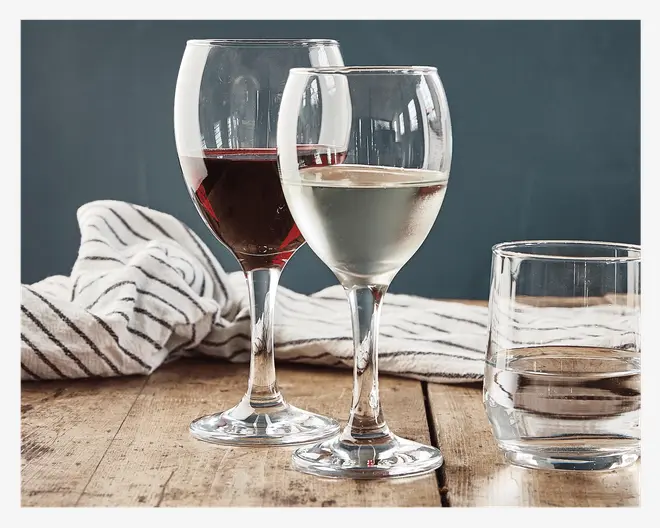 To klare vinglas, det ene fyldt med rødvin og det andet med hvidvin, står på et rustikt træbord. Ved siden af står et lavt, klart glas fyldt med vand. I baggrunden ligger en hvid klud med sorte striber, som giver et afslappet og elegant udtryk til opstillingen.