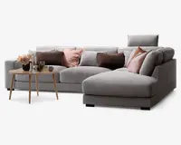 Moderne hjørnesofa betrukket med lysegråt stof, med chaiselong på højre side. Sofaen er pyntet med dekorative puder i bløde rosa, brune og grå nuancer, som giver ekstra komfort og stil. Foran sofaen står et rundt sofabord i lyst træ, dekoreret med en sort vase med blomster, stearinlys og små pyntegenstande. Sofaen har et rent, moderne design med brede armlæn og en støttende nakkestøtte i den ene ende.