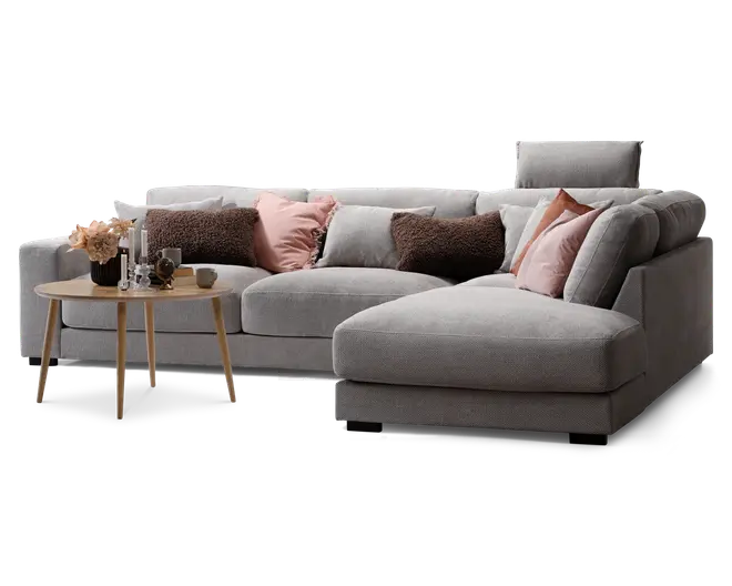 Moderne hjørnesofa betrukket med lysegråt stof, med chaiselong på højre side. Sofaen er pyntet med dekorative puder i bløde rosa, brune og grå nuancer, som giver ekstra komfort og stil. Foran sofaen står et rundt sofabord i lyst træ, dekoreret med en sort vase med blomster, stearinlys og små pyntegenstande. Sofaen har et rent, moderne design med brede armlæn og en støttende nakkestøtte i den ene ende.