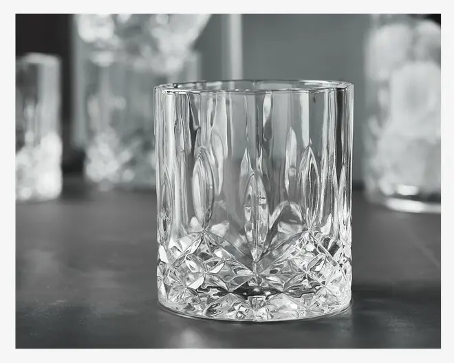 Klar glas tumbler med en tyk, tung bund, der har et slebet mønster af geometriske og bladformede figurer. Siderne er lige og glatte, mens den nederste halvdel er dekoreret med indviklede, facetslebne detaljer, der reflekterer lyset. Glasset har et robust og elegant udseende, velegnet til servering af spiritus eller cocktails.
