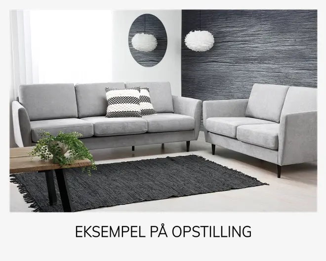 Et moderne stuearrangement med en lysegrå tresæders sofa og en matchende lænestol, begge med rene linjer og slanke, skrå sorte ben. Sofaen har to pyntepuder med sort-hvide geometriske mønstre. Rummet indeholder også et tekstureret mørkegråt tæppe, et sofabord i træ med en grøn plante og et rundt vægspejl. Bagvæggen har en tekstureret mørk overflade, og en hvid pendellampe med et luftigt udseende hænger fra loftet.