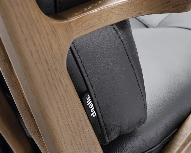 Nærbillede af en stol med en glat, sort læderpolstring med synlige syninger og et lille stofmærke med teksten 'daells'. Polstringen understøttes af en solid, lysebrun træramme med naturlig træstruktur. Billedet fremhæver materialernes tekstur og kvalitet i stolens konstruktion.