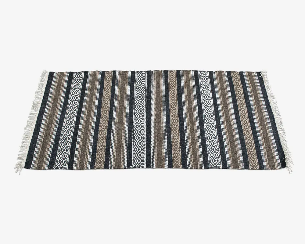 Rektangulært vævet tekstil med skiftende lodrette striber i sorte, brune og beige nuancer, hvor nogle striber har indviklede geometriske mønstre. Kanterne er afsluttet med hvide frynser på begge korte sider.