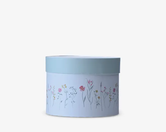 En lyseblå cylindrisk beholder med matchende låg, dekoreret hele vejen rundt med et fint blomstermønster i bløde rosa, gule og grå nuancer.