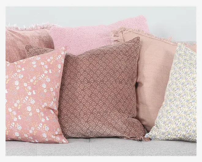 En samling pyntepuder i forskellige pastelfarver og mønstre. Puderne inkluderer en lyserød med små blomster, en brun med et diskret geometrisk mønster, en ensfarvet lyserød pude med frynser i kanten, en blød, fluffy lyserød pude og en pude med et sart blåt og gult blomstermønster. Teksturerne varierer fra glat hør til blødt, plysset stof, og det samlede udtryk er hyggeligt og indbydende.