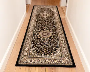 En lang, rektangulær løber med et traditionelt persisk-inspireret design. Tæppet har en mørk bundfarve med indviklede blomster- og rankemønstre i beige, creme og bordeaux nuancer. Der er to store medaljonmotiver placeret langs midten, og kanten er dekoreret med detaljerede, gentagne blomstermønstre i lyse farver. Tæppet har en lav luv og ser ud til at være lavet af et tæt vævet materiale.