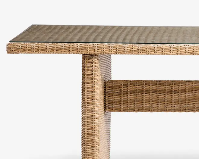 Nærbillede af et møbel med flettet rattan- eller fletstruktur i en naturlig lysebrun farve. Overfladen er dækket af en klar glasplade, som giver en glat og holdbar finish. Konstruktionen har en lodret støtte og en vandret tværstiver, begge med samme flettede design, hvilket giver et solidt og stilfuldt udtryk, der egner sig til både indendørs og udendørs brug.