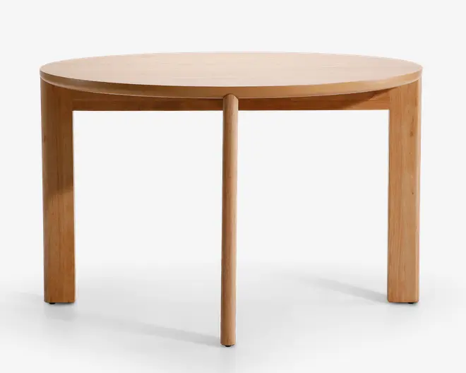 Rundt bord i lyst træ med en glat overflade. Bordet har et minimalistisk design med tre lige ben og et centralt, afrundet ben, der giver både stabilitet og et unikt visuelt udtryk. Kanterne er blødt buede, og det samlede udtryk er moderne og enkelt.