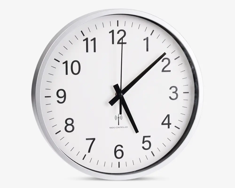 Rund vægur med hvid urskive, sorte time- og minutvisere samt en tynd sort sekundviser. Uret har store, tydelige sorte arabiske tal for hver time og minutmarkeringer langs kanten. Det har en enkel, moderne sølvramme og klart glas foran. Klokken viser 5:08:00. Under midten er der et lille symbol og tekst, der indikerer, at uret er radiostyret for præcis tidsangivelse.