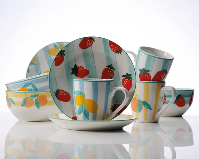 Et sæt keramikservice med livlige, håndmalede frugtmotiver. Sættet består af tallerkener, skåle og krus, alle dekoreret med farverige illustrationer af jordbær, citroner og appelsiner. Baggrunden på delene veksler mellem pastelfarvede blå og gule lodrette striber, hvilket giver et legende og frisk udtryk. Krusene har store, behagelige hanke, og den samlede stil er glad og moderne – perfekt til at skabe en hyggelig stemning ved bordet.