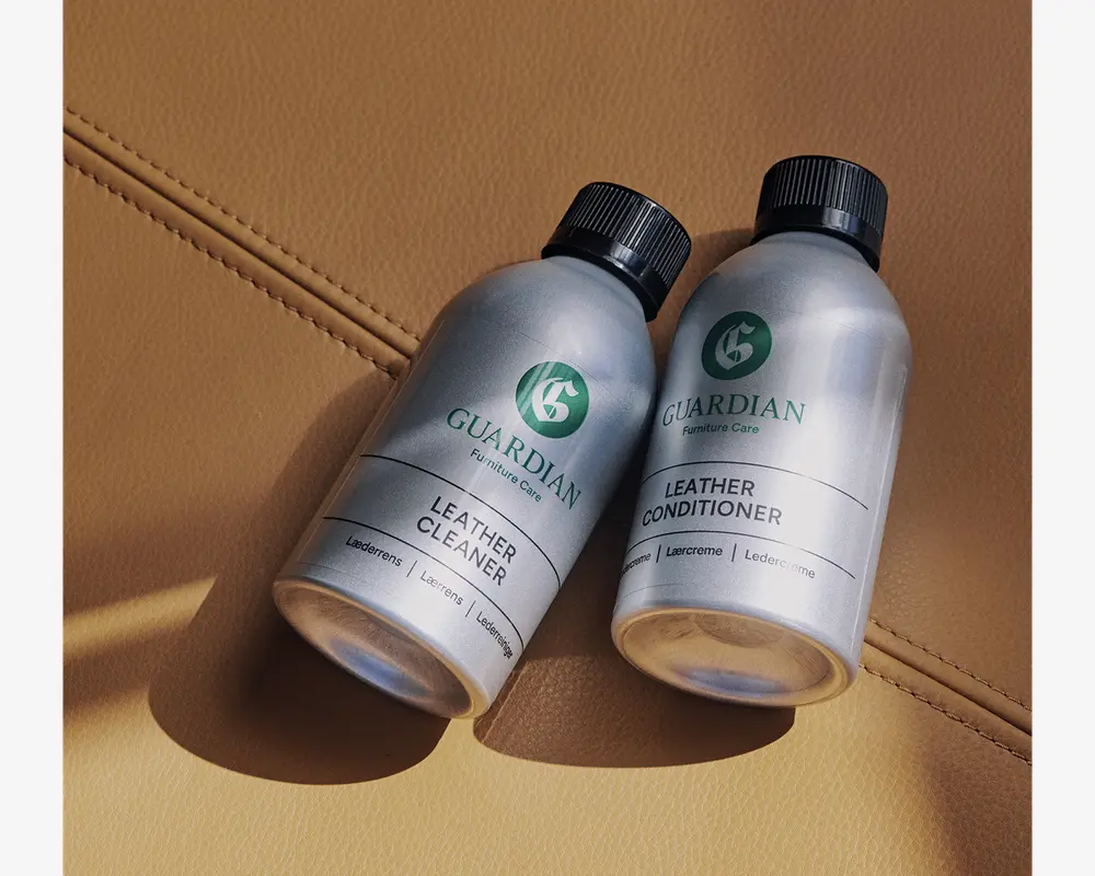 To sølvfarvede flasker med sorte låg vises, mærket 'Guardian Furniture Care.' Den ene flaske er mærket 'Leather Cleaner' og den anden 'Leather Conditioner.' Begge flasker har et minimalistisk design med grøn og sort tekst og ligger på en lysebrun læderoverflade med synlige syninger. Flaskerne ser ud til at være lavet af metal og er kompakte, hvilket gør dem nemme at håndtere og opbevare.