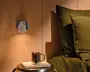 Moderne vægmonteret lampe med rund bagplade og kort cylindrisk skærm i mat lysegrå, som kaster et varmt nedadrettet lys på en væg med træpaneler.
