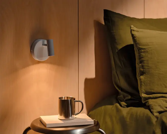 Moderne vægmonteret lampe med rund bagplade og kort cylindrisk skærm i mat lysegrå, som kaster et varmt nedadrettet lys på en væg med træpaneler.
